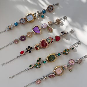 May include: Several decorative wristwatches with diverse face shapes and embellishments. The watches have silver-toned chains and are decorated with floral, heart, and gemstone accents. The watch faces display colours like purple, green, and pink.