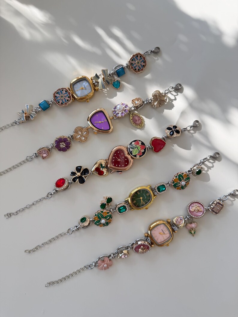 May include: Several decorative wristwatches with diverse face shapes and embellishments. The watches have silver-toned chains and are decorated with floral, heart, and gemstone accents. The watch faces display colours like purple, green, and pink.