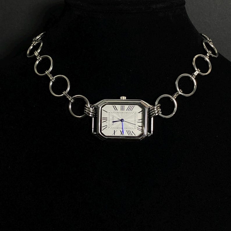 May include: A silver chain necklace with a rectangular watch face attached. The watch face has Roman numeral markings and a blue hand.
