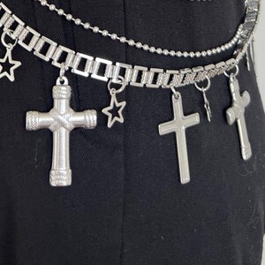 May include: Silver-toned belt with multiple cross and star charms. The belt has three strands: a chain of rectangular links, a beaded chain, and a chain of small links. The belt is shown against a black background.