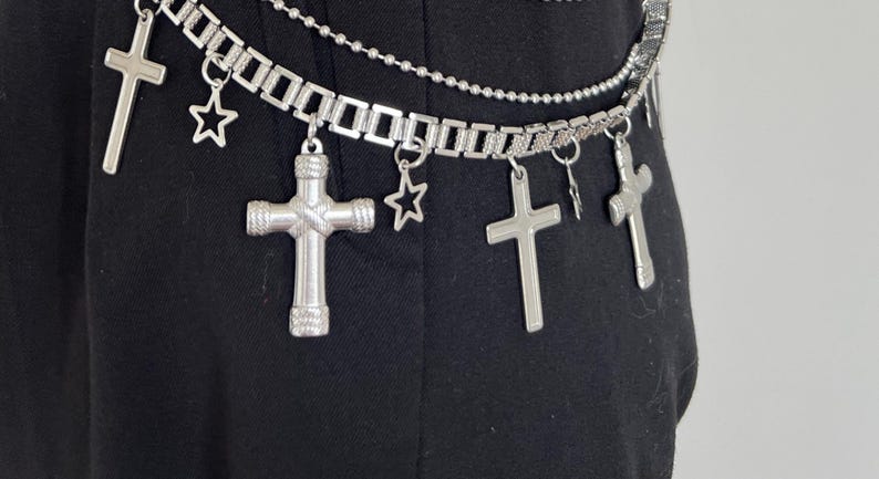 May include: Silver-toned belt with multiple cross and star charms. The belt has three strands: a chain of rectangular links, a beaded chain, and a chain of small links. The belt is shown against a black background.
