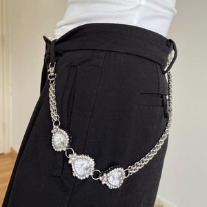 May include: A silver-toned chain belt with three heart-shaped watch faces. Each watch face is surrounded by small, clear crystals. The belt is attached to black pants.