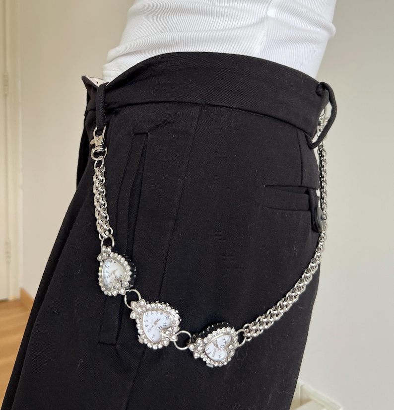 May include: A silver-toned chain belt with three heart-shaped watch faces. Each watch face is surrounded by small, clear crystals. The belt is attached to black pants.