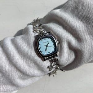 May include: A silver-toned wristwatch with a square, light blue face. The watch features the word "OURSTART" and has a delicate, floral-patterned band. The watch is displayed on a white surface.