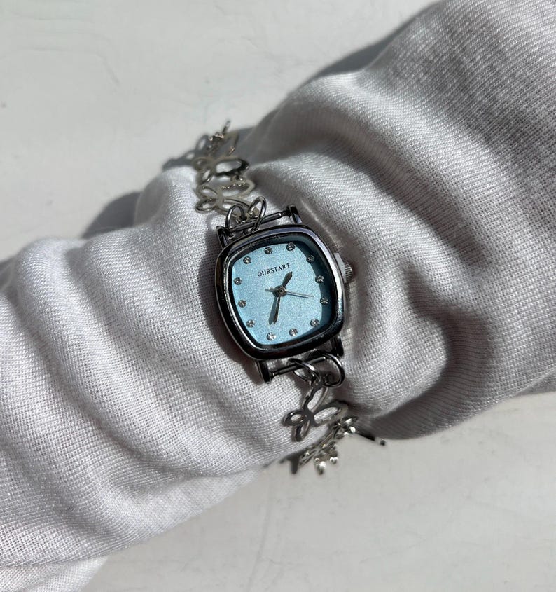 May include: A silver-toned wristwatch with a square, light blue face. The watch features the word "OURSTART" and has a delicate, floral-patterned band. The watch is displayed on a white surface.