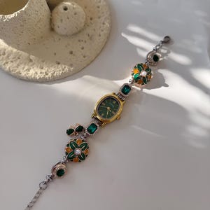 May include: A gold wristwatch with a green face and decorative green and gold floral accents. The watch band features a silver chain and green gemstone embellishments. The watch is displayed on a white surface.