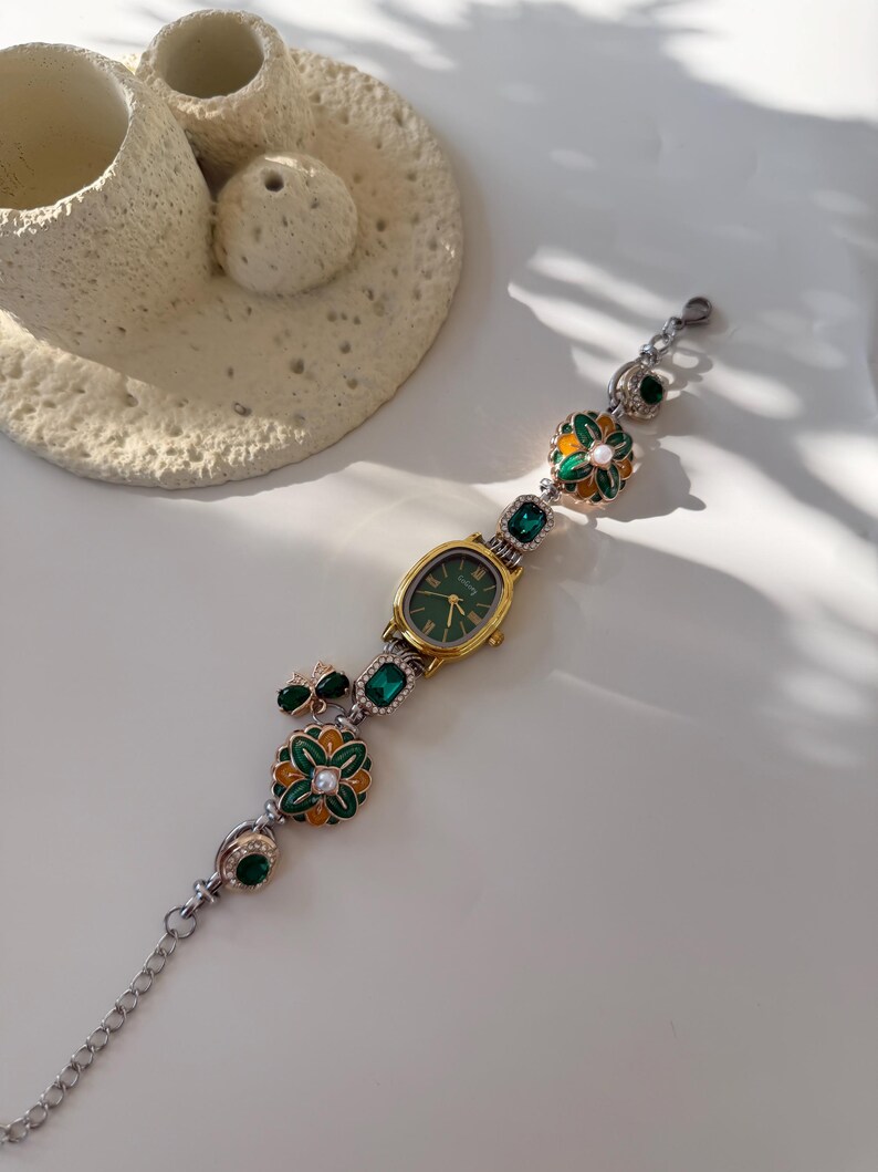 May include: A gold wristwatch with a green face and decorative green and gold floral accents. The watch band features a silver chain and green gemstone embellishments. The watch is displayed on a white surface.