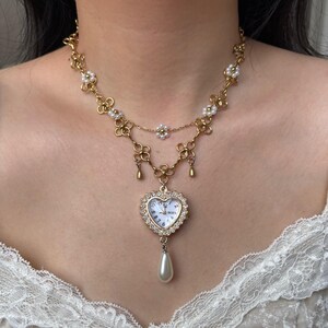 Puede incluir: Un collar dorado con un colgante de reloj en forma de coraz&oacute;n y una gota de perla. El collar presenta un dise&ntilde;o floral con peque&ntilde;as perlas y una cadena delicada. La esfera del reloj muestra la hora. El collar se lleva alrededor del cuello.