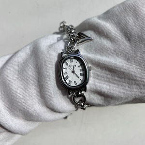 May include: Silver-toned wristwatch with an oval face and Roman numeral hour markers. The watch has a chain-link bracelet and a small heart-shaped charm. The watch face is white with the brand name "Bogory".