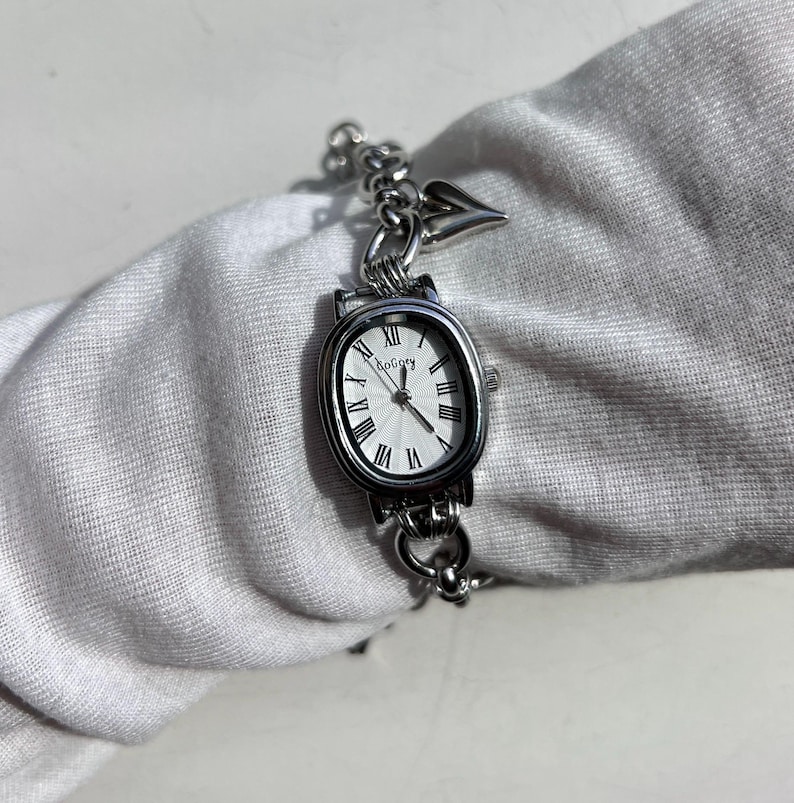 May include: Silver-toned wristwatch with an oval face and Roman numeral hour markers. The watch has a chain-link bracelet and a small heart-shaped charm. The watch face is white with the brand name "Bogory".