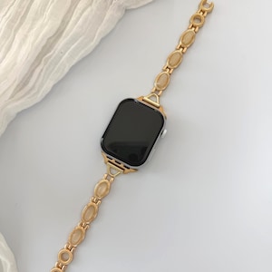 May include: Gold-toned watch band with oval-shaped, light-colored stones. The band is attached to a black-screened smartwatch. The watch band is made of a gold-colored metal and has a delicate, elegant design.