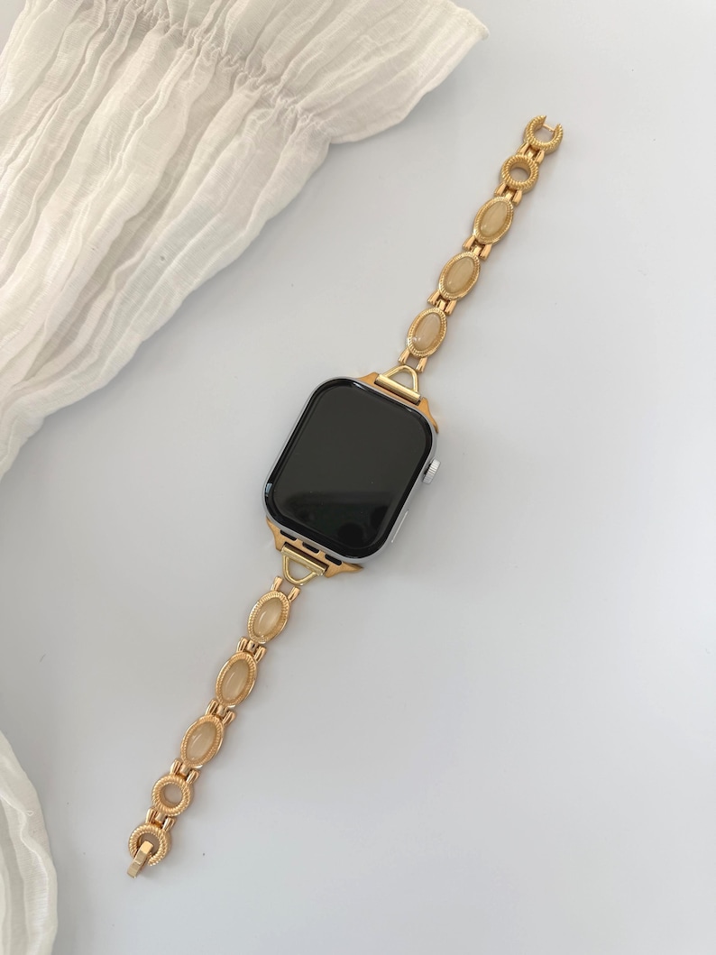 May include: Gold-toned watch band with oval-shaped, light-colored stones. The band is attached to a black-screened smartwatch. The watch band is made of a gold-colored metal and has a delicate, elegant design.