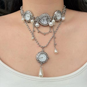 Op de afbeelding: Zilverkleurige choker-ketting met meerdere horlogezichten en imitatieparels. De ketting heeft een hartvormig horlogezicht in het midden, met ronde horlogezichten aan weerszijden. Hangende kettingen en een druppelvormige parelhanger voegen details toe.