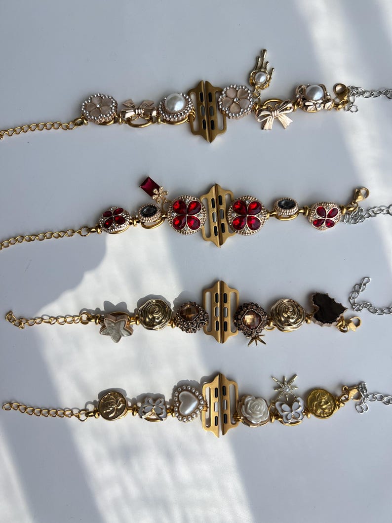 May include: Four decorative watch bands with gold and silver chains. Each band features a gold-colored central connector and is adorned with various embellishments, including pearls, red gems, and floral designs. The bands are displayed on a white surface.