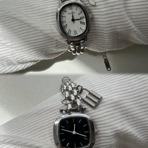 May include: Two silver watches with chain link bands and charms. The top watch has a white face with Roman numeral hour markers and a silver charm shaped like a cross. The bottom watch has a black face with white hour and minute hands and a silver padlock charm.