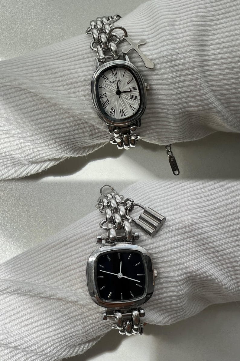 May include: Two silver watches with chain link bands and charms. The top watch has a white face with Roman numeral hour markers and a silver charm shaped like a cross. The bottom watch has a black face with white hour and minute hands and a silver padlock charm.