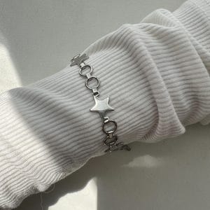 May include: A silver-toned chain bracelet featuring alternating star-shaped charms and circular metal links. The bracelet is displayed on a white ribbed fabric sleeve, showcasing a metallic finish and a repeating pattern of stars and rings.