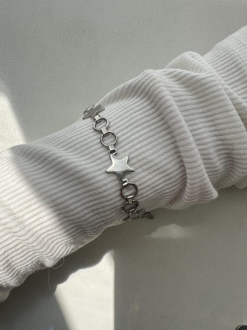 May include: A silver-toned chain bracelet featuring alternating star-shaped charms and circular metal links. The bracelet is displayed on a white ribbed fabric sleeve, showcasing a metallic finish and a repeating pattern of stars and rings.
