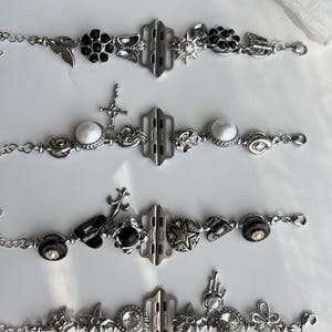 May include: Four silver-toned bracelets with various decorative charms. Charms include black flowers, butterflies, stars, crosses, and round white stones. Each bracelet has a central rectangular clasp and a chain link band.