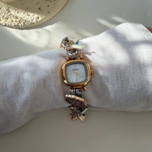 May include: A gold-toned wristwatch with a square face and a light blue dial. The watch band features decorative metal links in gold and silver tones. The watch face reads "LEAEK".