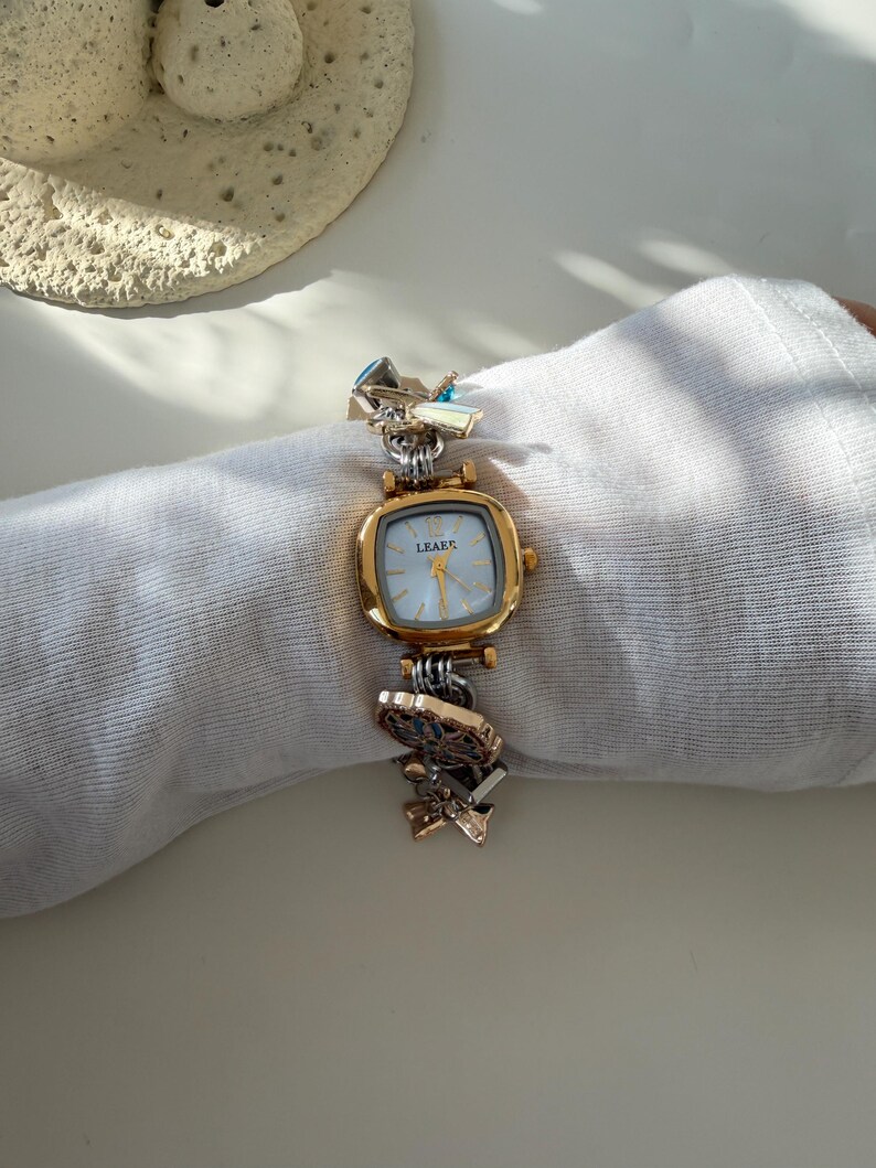 May include: A gold-toned wristwatch with a square face and a light blue dial. The watch band features decorative metal links in gold and silver tones. The watch face reads "LEAEK".