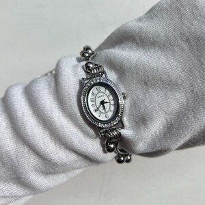 May include: Silver-toned wristwatch with an oval face and a white dial. The watch features a crystal-studded bezel and a delicate, ornate band. The watch face displays the brand name "NAIDU" and traditional hour markers.