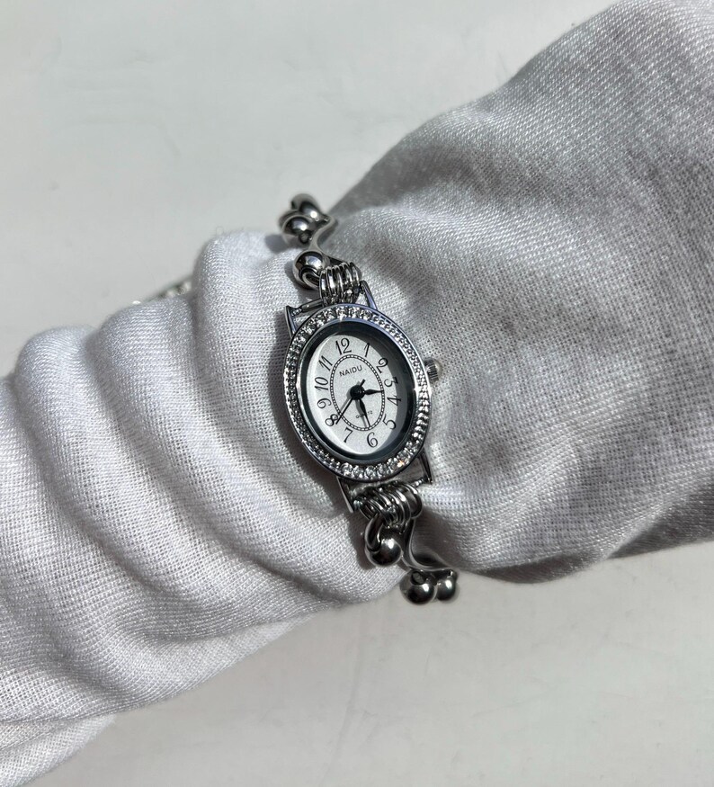 May include: Silver-toned wristwatch with an oval face and a white dial. The watch features a crystal-studded bezel and a delicate, ornate band. The watch face displays the brand name "NAIDU" and traditional hour markers.