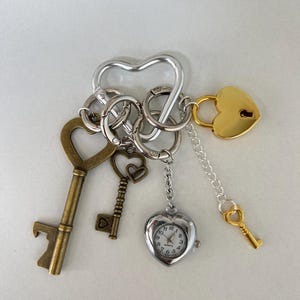 Puede incluir: Un llavero de corazón plateado con múltiples dijes, incluyendo un candado de corazón dorado, dos llaves de estilo antiguo, un pequeño dije de corazón plateado y un reloj de corazón plateado.