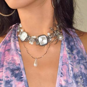May include: A silver choker necklace with two square watches, a pearl drop pendant, and a silver spider charm. The necklace is on a person wearing a purple and blue tie-dye shirt.