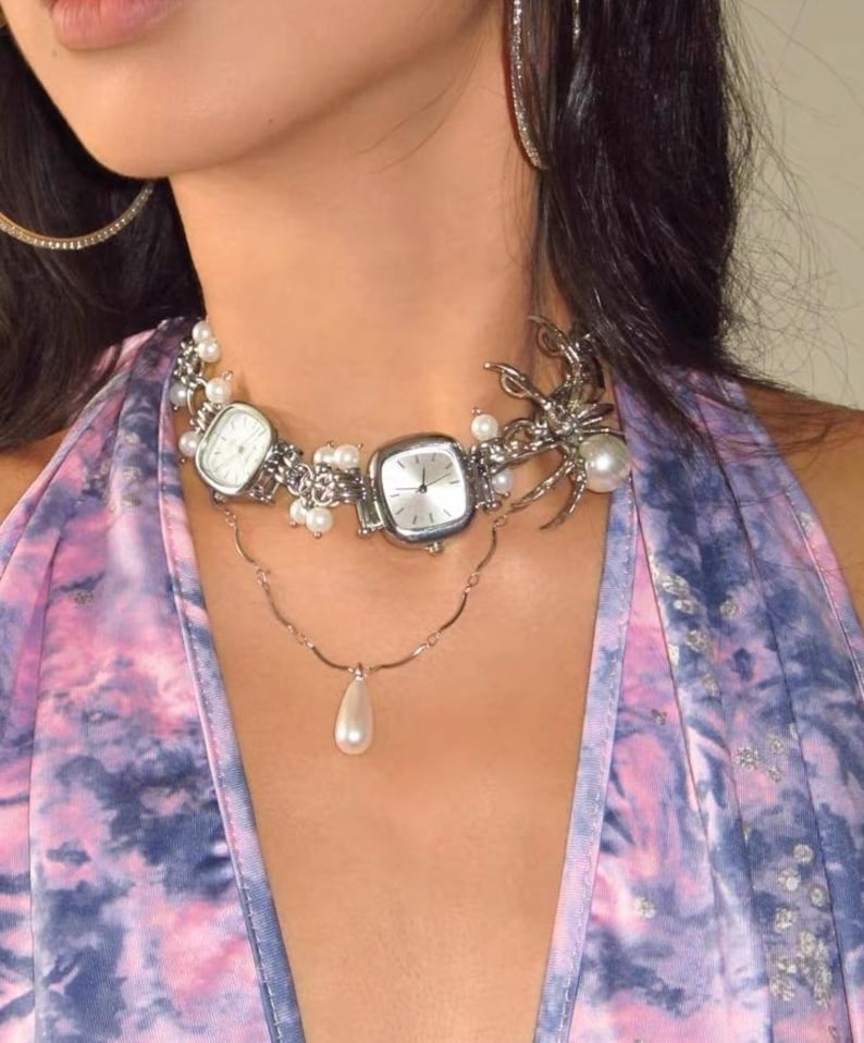 May include: A silver choker necklace with two square watches, a pearl drop pendant, and a silver spider charm. The necklace is on a person wearing a purple and blue tie-dye shirt.