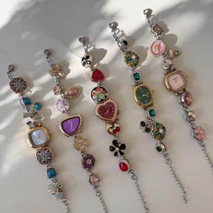 May include: Five decorative charm bracelets with silver chains and various colourful charms. Each bracelet features a small watch face with a different coloured dial. Charms include flowers, hearts, and gemstones.