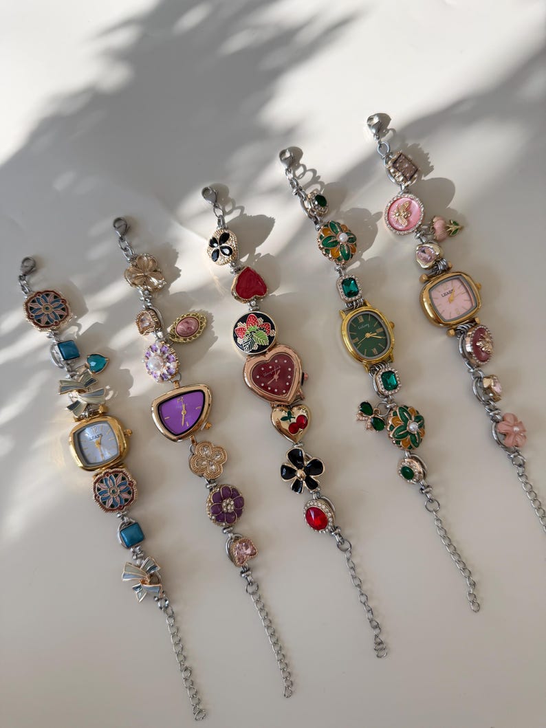 May include: Five decorative charm bracelets with silver chains and various colourful charms. Each bracelet features a small watch face with a different coloured dial. Charms include flowers, hearts, and gemstones.