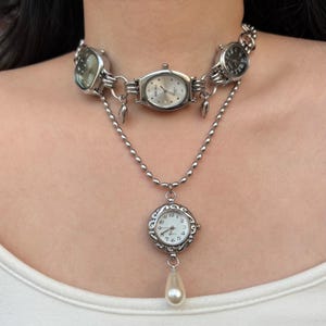 Könnte beinhalten: Silberfarbene Uhrenkette mit mehreren Zifferblättern und einem Perlenanhänger. Die Kette hat ein mehrschichtiges Design mit kleinen Zifferblättern, die das Halsband bilden, und einem größeren Zifferblatt mit einem Perlentropfen. Die Zifferblätter sind silber und schwarz.