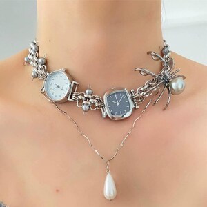 May include: A silver necklace with three silver watches and a silver spider charm with a pearl. The necklace also has a pearl pendant.