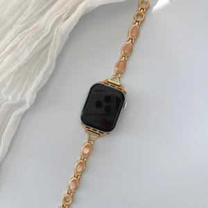 May include: Gold-toned Apple Watch band with a chain-link design. Each link features an oval-shaped, peach-colored stone. The watch face is black, and the band is set against a white background with a soft, draped fabric.