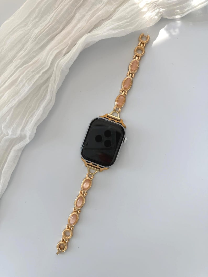 May include: Gold-toned Apple Watch band with a chain-link design. Each link features an oval-shaped, peach-colored stone. The watch face is black, and the band is set against a white background with a soft, draped fabric.