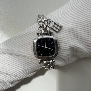 May include: A silver-toned analog wristwatch featuring a square-shaped case with rounded corners and a black dial. The watch has a multi-link chain bracelet and includes a small, dangling silver-toned padlock charm attached to the watch band.