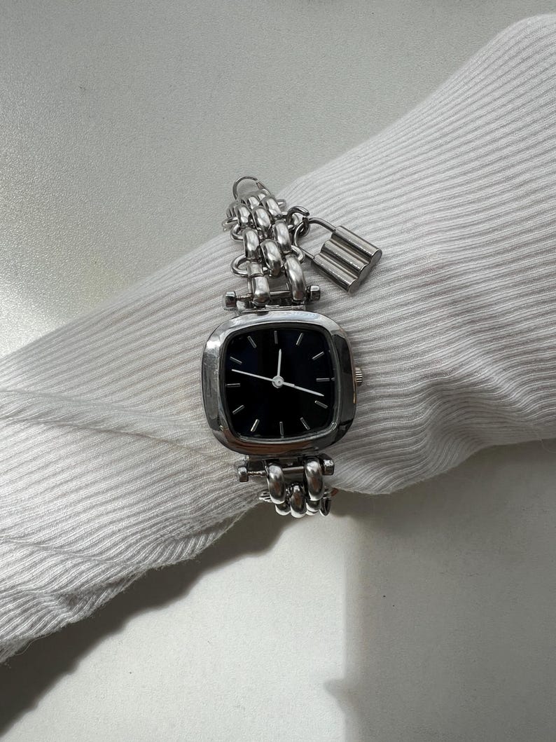 May include: A silver-toned analog wristwatch featuring a square-shaped case with rounded corners and a black dial. The watch has a multi-link chain bracelet and includes a small, dangling silver-toned padlock charm attached to the watch band.