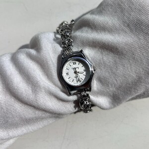 May include: A silver-toned wristwatch with a white face and black Roman numerals. The watch has a decorative band and the word "Gallery" is printed on the watch face. The watch is a quartz movement.