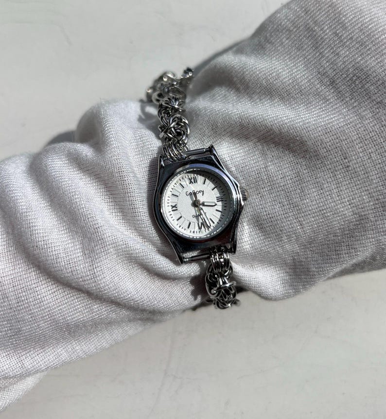 May include: A silver-toned wristwatch with a white face and black Roman numerals. The watch has a decorative band and the word "Gallery" is printed on the watch face. The watch is a quartz movement.