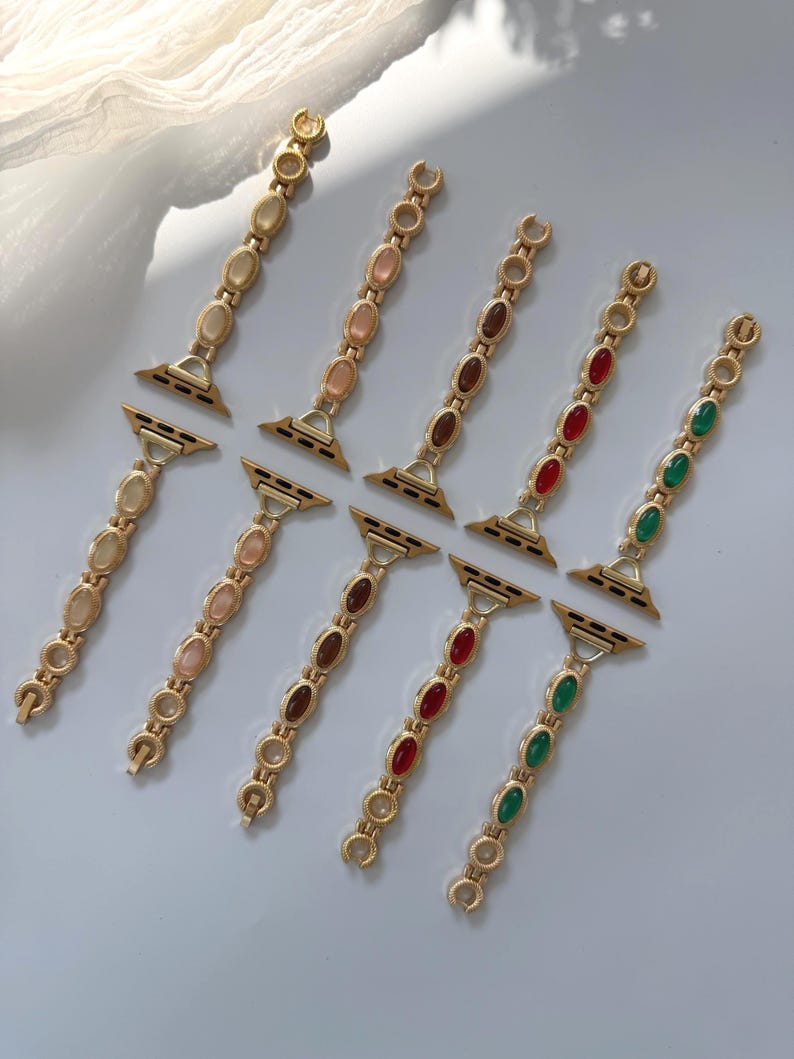 May include: Gold-toned watch bands with oval-shaped gemstones in various colors, including cream, pink, brown, red, and green. The bands are designed with a chain-like structure and are displayed on a light-colored surface.