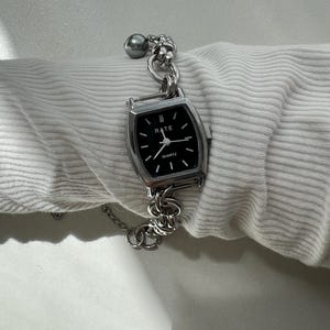 May include: A silver-toned quartz wristwatch with a rectangular black face featuring the text RATE and QUARTZ. The watch has a thick, chunky silver-colored chain link bracelet and a single grey spherical bead accent attached to the metal links.