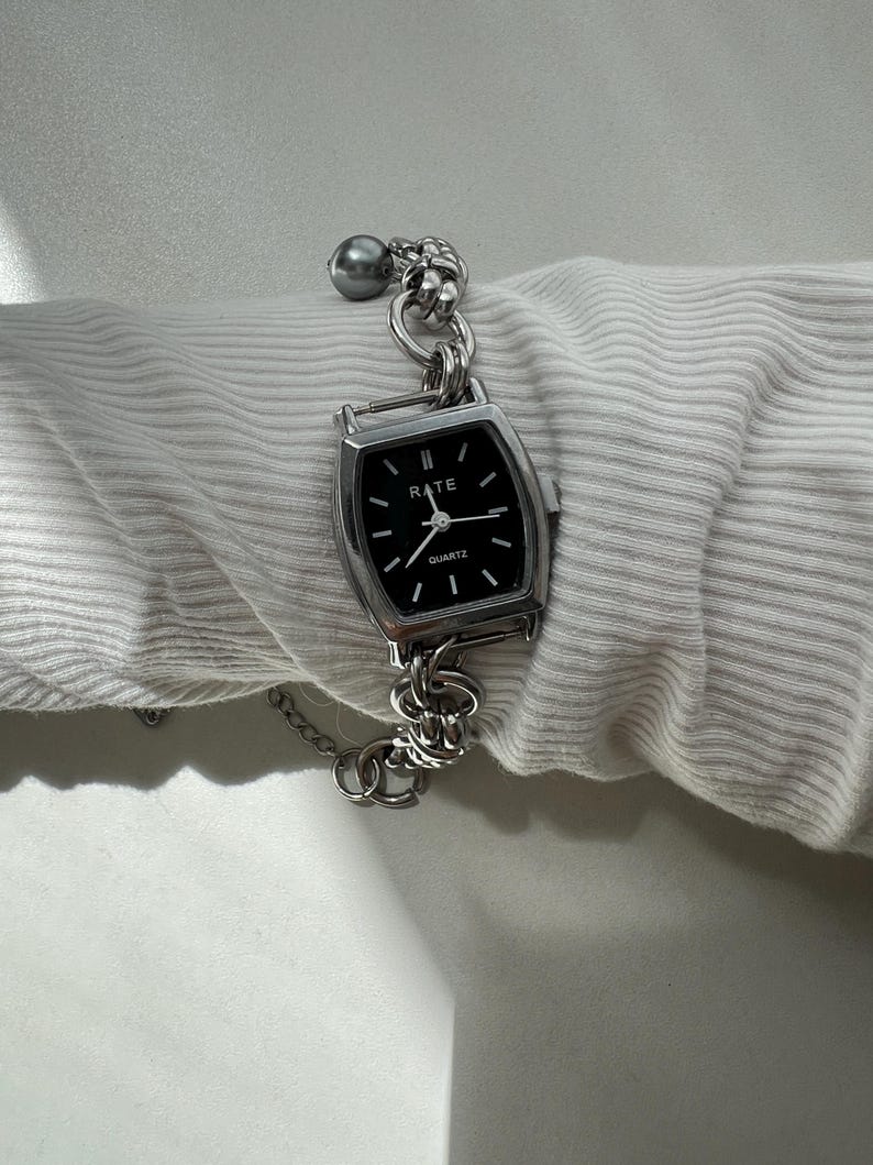 May include: A silver-toned quartz wristwatch with a rectangular black face featuring the text RATE and QUARTZ. The watch has a thick, chunky silver-colored chain link bracelet and a single grey spherical bead accent attached to the metal links.