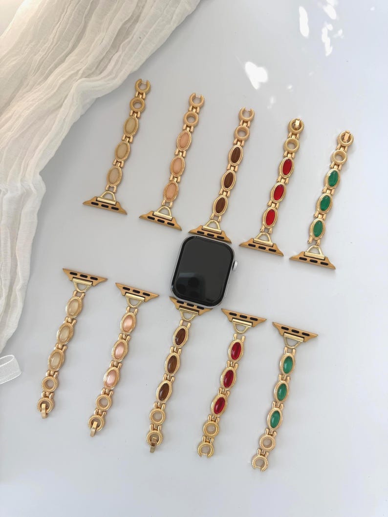 May include: Gold-toned watch bands with oval-shaped colored accents. The bands are in various colors, including beige, brown, red, and green. The bands are designed to fit a smartwatch.