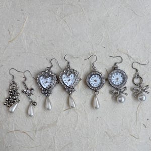 May include: Set of six silver earrings with pearl accents.  Two pairs feature heart-shaped watch faces, two pairs feature round watch faces, and one pair features a bow and a circle.