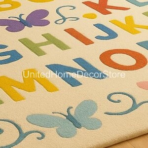 K&ouml;nnte beinhalten: Nahaufnahme eines cremefarbenen Teppichs mit bunten Buchstaben und Schmetterlingsmotiven. Die Buchstaben sind in verschiedenen Farben gehalten, darunter Blau, Orange, Gelb und Gr&uuml;n. Der Teppich tr&auml;gt den Text "United Home Decor Store".