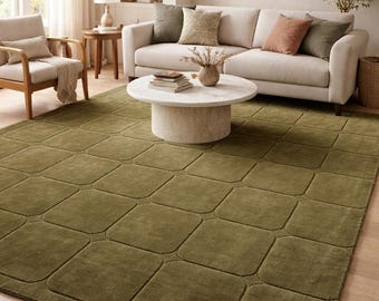 May include: A large olive green area rug with a geometric pattern dominates the foreground of a living room. The rug's design features a series of connected squares. A beige sofa with decorative pillows and a round coffee table are also visible.