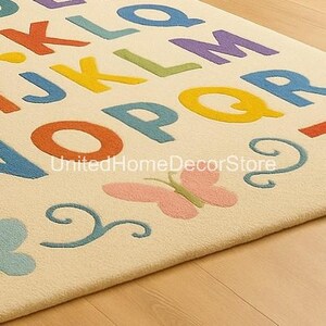 K&ouml;nnte beinhalten: Cremefarbener Teppich mit bunten Buchstaben und einem rosa Schmetterlingsdesign. Die Buchstaben sind in verschiedenen Farben, darunter Orange, Blau, Gelb und Gr&uuml;n. Der Teppich hat ein weiches, strukturiertes Aussehen.