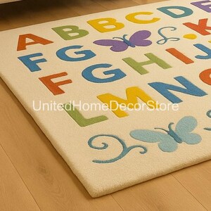 K&ouml;nnte beinhalten: Cremefarbener Teppich mit dem Alphabet in verschiedenen Farben, Schmetterlingen und Wirbeldesigns. Der Teppich tr&auml;gt den Text "UnitedHomeDecorStore". Der Teppich liegt auf einem Holzboden.
