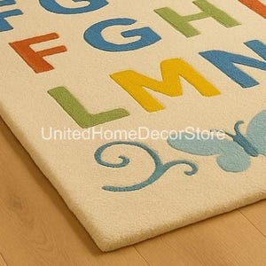 K&ouml;nnte beinhalten: Cremefarbener Teppich mit bunten Buchstaben des Alphabets. Die Buchstaben sind in Orange, Gr&uuml;n, Blau und Gelb gehalten. Ein blaues Schmetterlingsdesign befindet sich in der Ecke. Der Teppich liegt auf einem Holzboden.